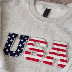 May include: A gray sweatshirt with a large "USA" graphic on the front. The letters are made of red, white, and blue fabric with a silver glitter outline.