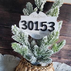 Puede incluir: Adorno navideño blanco con texto negro que dice "Christmas 30153 Rockmart, Georgia 2022". El adorno está colgado de un pequeño pino artificial con nieve blanca.
