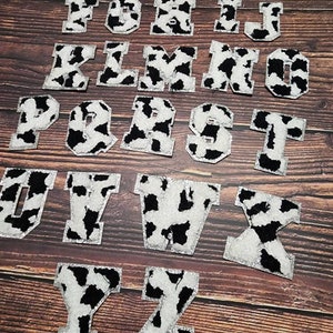 Peut inclure: Un ensemble de 26 lettres thermocollantes &agrave; motif vache. Les lettres sont blanches avec des taches noires et ont une finition paillet&eacute;e. Les lettres sont dispos&eacute;es en ordre alphab&eacute;tique sur une surface en bois.