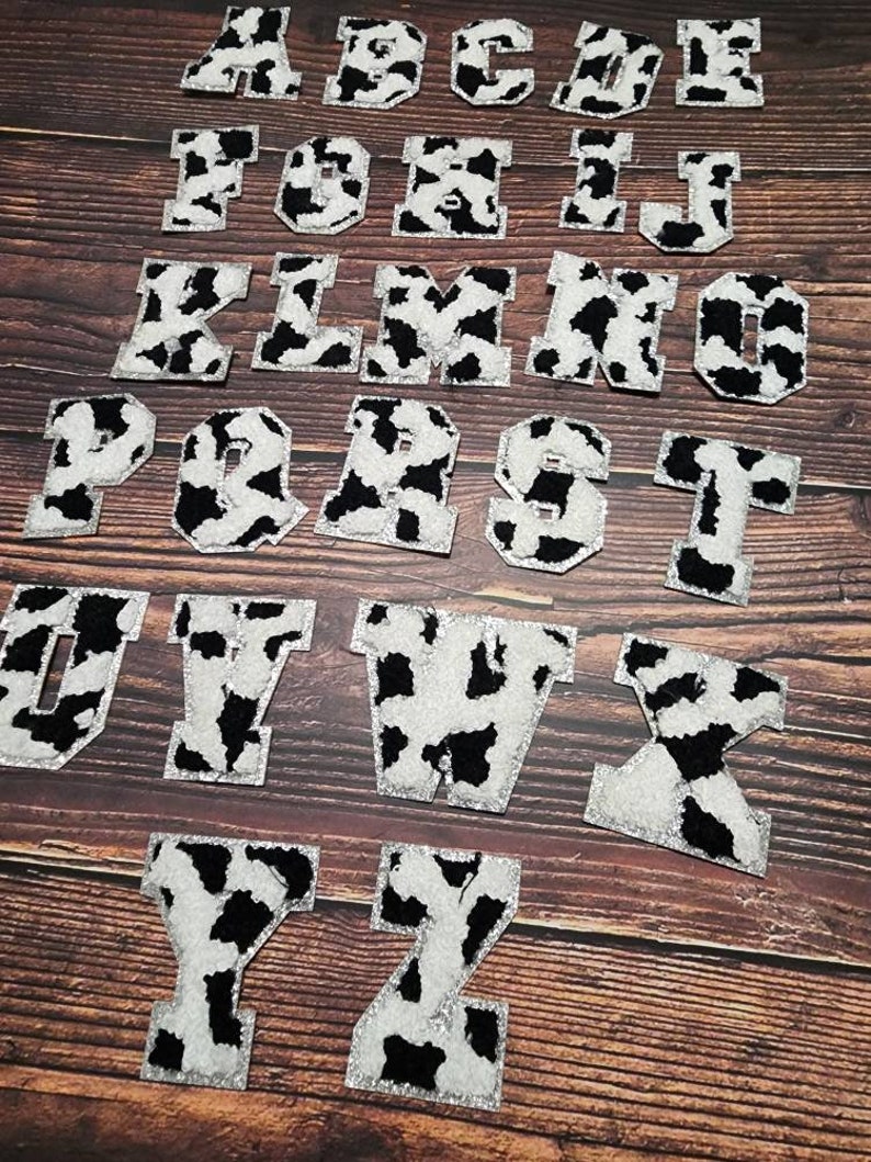 Peut inclure: Un ensemble de 26 lettres thermocollantes &agrave; motif vache. Les lettres sont blanches avec des taches noires et ont une finition paillet&eacute;e. Les lettres sont dispos&eacute;es en ordre alphab&eacute;tique sur une surface en bois.