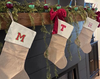 May include: Christmas stockings with embroidered letters hang above a fireplace, adorned with a red velvet bow and vintage Christmas lights. The stockings are beige with white tops and green and red embroidery.