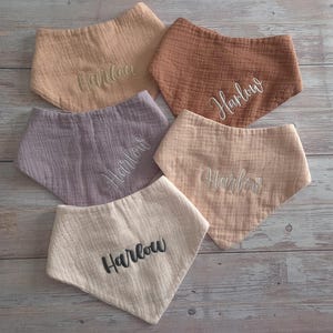 May include: Five triangular bibs made of textured muslin fabric in shades of beige, tan, brown, and dusty purple. Each bib features the name Harlow embroidered in a script font in either white, dark gray, or matching thread colors.