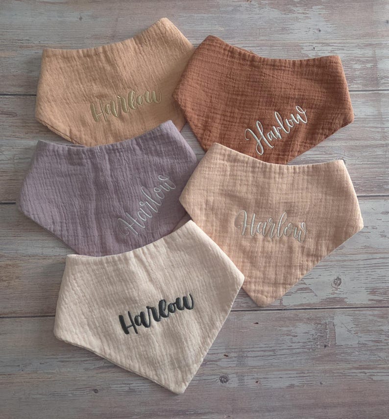 May include: Five triangular bibs made of textured muslin fabric in shades of beige, tan, brown, and dusty purple. Each bib features the name Harlow embroidered in a script font in either white, dark gray, or matching thread colors.