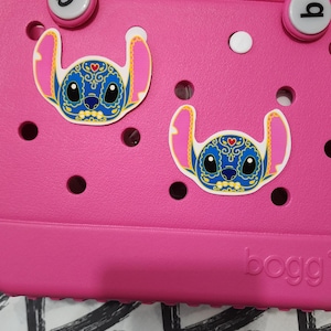 May include: Pink plastic tote bag with two blue and white Stitch character stickers with a Day of the Dead design. The bag has a white circle and the word "bogg" on the bottom.