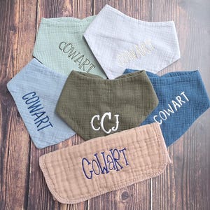 May include: A collection of six muslin fabric bibs in various colors including sage green, white, light blue, olive green, dark blue, and dusty rose. Each bib features embroidered text, with five displaying the name COWART and one showing CCJ.