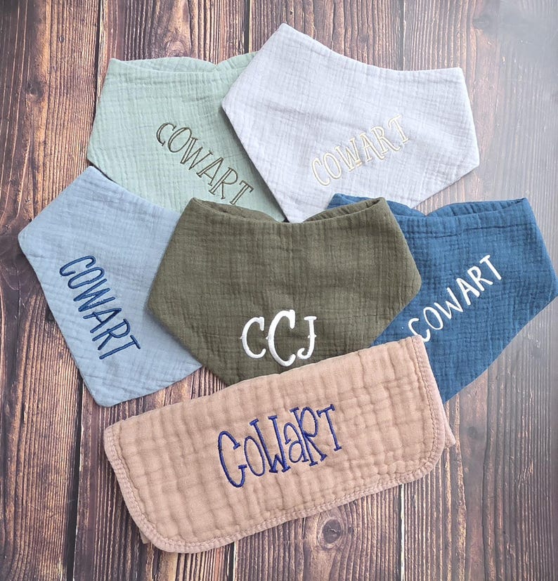May include: A collection of six muslin fabric bibs in various colors including sage green, white, light blue, olive green, dark blue, and dusty rose. Each bib features embroidered text, with five displaying the name COWART and one showing CCJ.