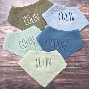 May include: Five triangular muslin baby bibs in olive green, light blue, sage green, dark blue, and pale green. Each bib features embroidered names including Colin or Colin Charles in white, dark blue, or olive green thread across the center.