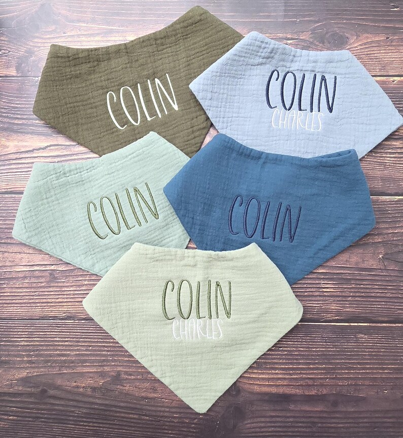 May include: Five triangular muslin baby bibs in olive green, light blue, sage green, dark blue, and pale green. Each bib features embroidered names including Colin or Colin Charles in white, dark blue, or olive green thread across the center.