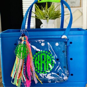 May include: A bright blue tote bag with a clear zippered pouch on the front. The pouch has a green monogram with white polka dots that says "KRR". The bag has a colorful tassel with a beaded charm attached to the handle.