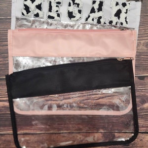 Peut inclure: Trois pochettes transparentes &agrave; fermeture &eacute;clair avec une doublure en tissu noir, rose et blanc. La pochette sup&eacute;rieure a une doublure en tissu blanc avec le mot "MEGAN" en lettres d'impression vache noire et blanche.