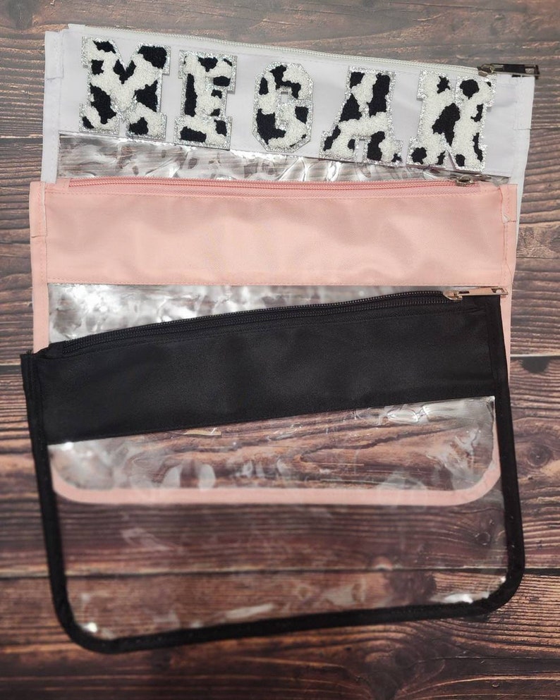 Peut inclure: Trois pochettes transparentes &agrave; fermeture &eacute;clair avec une doublure en tissu noir, rose et blanc. La pochette sup&eacute;rieure a une doublure en tissu blanc avec le mot "MEGAN" en lettres d'impression vache noire et blanche.