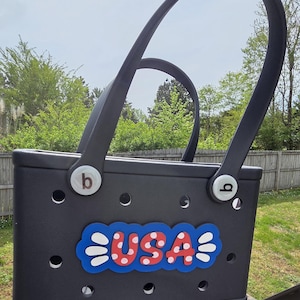 May include: A black tote bag with a USA design. The bag has two curved handles and a decorative USA emblem in red, white, and blue. The bag has a series of small holes around the body.