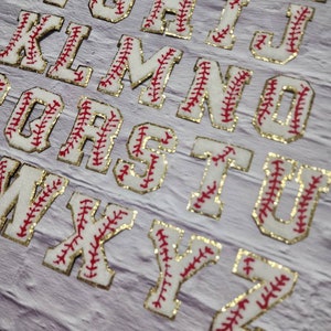 以下が含まれることがあります： 赤と金色のグリッタートリムで野球のステッチデザインが施された白いフェルトのアルファベット文字