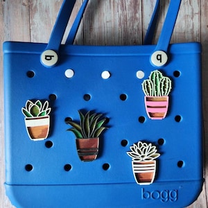 May include: Blue Bogg bag with white dots and four different plant-themed charms. The charms are a cactus, a succulent, and two different types of leafy plants. The charms are attached to the bag with a white button.