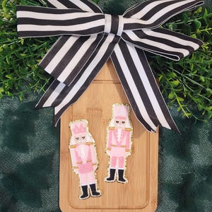 Könnte beinhalten: Zwei rosa-goldene Nussknacker-Ornamente auf einem Holzbrett, gekrönt von einer schwarz-weiß gestreiften Schleife. Die Ornamente haben schwarze Stiefel und sind von grünem Laub umgeben.