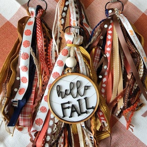 May include: Three autumn-themed keychains with a variety of ribbons and embellishments. The centre keychain features a round wooden sign with the text "hello fall".