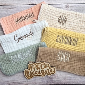 May include: A collection of personalized baby burp cloths in various colors, including peach, white, yellow, and sage green. Each cloth is embroidered with a name or initials. The wooden sign in the image reads "H&Co Designs."