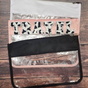 Peut inclure: Une pochette zipp&eacute;e en plastique transparent avec une fermeture &eacute;clair noire et une doublure en tissu rose. La pochette est orn&eacute;e d'un patch en tissu imprim&eacute; vache noir et blanc avec l'inscription "TRAVEL".