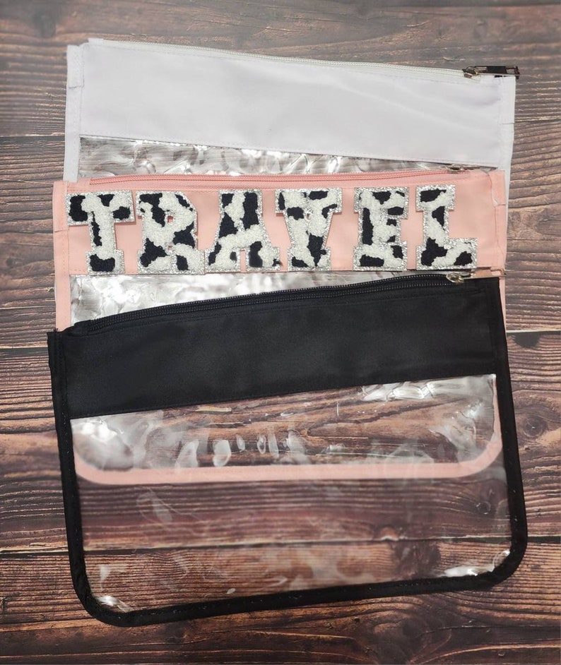 Peut inclure: Une pochette zipp&eacute;e en plastique transparent avec une fermeture &eacute;clair noire et une doublure en tissu rose. La pochette est orn&eacute;e d'un patch en tissu imprim&eacute; vache noir et blanc avec l'inscription "TRAVEL".