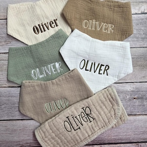 May include: A collection of six muslin baby bibs in various neutral colors, including beige, brown, green, and white. Each bib is embroidered with the name "Oliver" in a coordinating thread color.