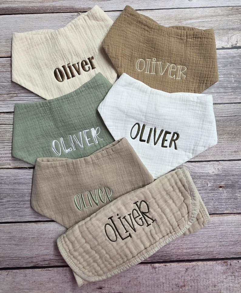 May include: Six triangular muslin bibs in various neutral shades including cream, sage green, and tan. Each bib features the name oliver embroidered in different fonts and thread colors, such as dark brown, white, and light green, across the center.