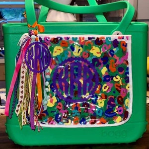 May include: Green tote bag with a clear plastic pocket featuring a colorful abstract print and a purple monogram. The bag has a white zipper and a white label that says "baggallini".