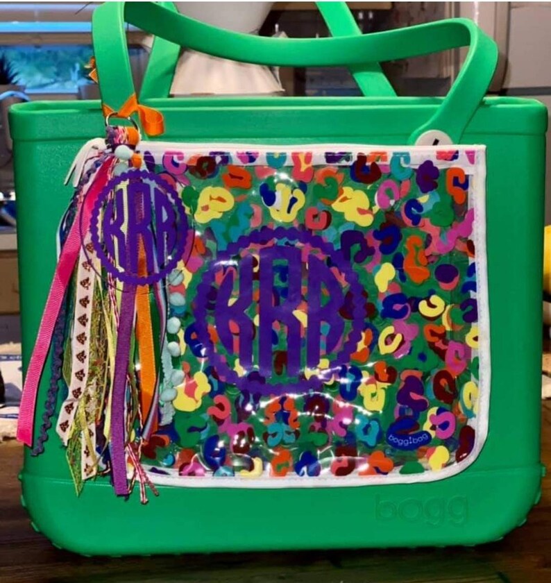 May include: Green tote bag with a clear plastic pocket featuring a colorful abstract print and a purple monogram. The bag has a white zipper and a white label that says "baggallini".