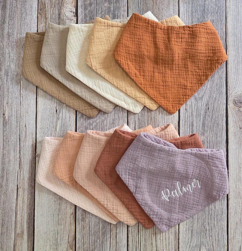 May include: A collection of triangular bibs made from textured muslin fabric in various earth tones including tan, cream, rust, peach, and mauve. One mauve bib features the name Palmer embroidered in white cursive script on the front surface.