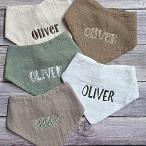 May include: Five triangular baby bibs made of textured muslin fabric in cream, tan, sage green, and white. Each bib features the name Oliver embroidered in the center in various thread colors including dark brown, light tan, white, and green.