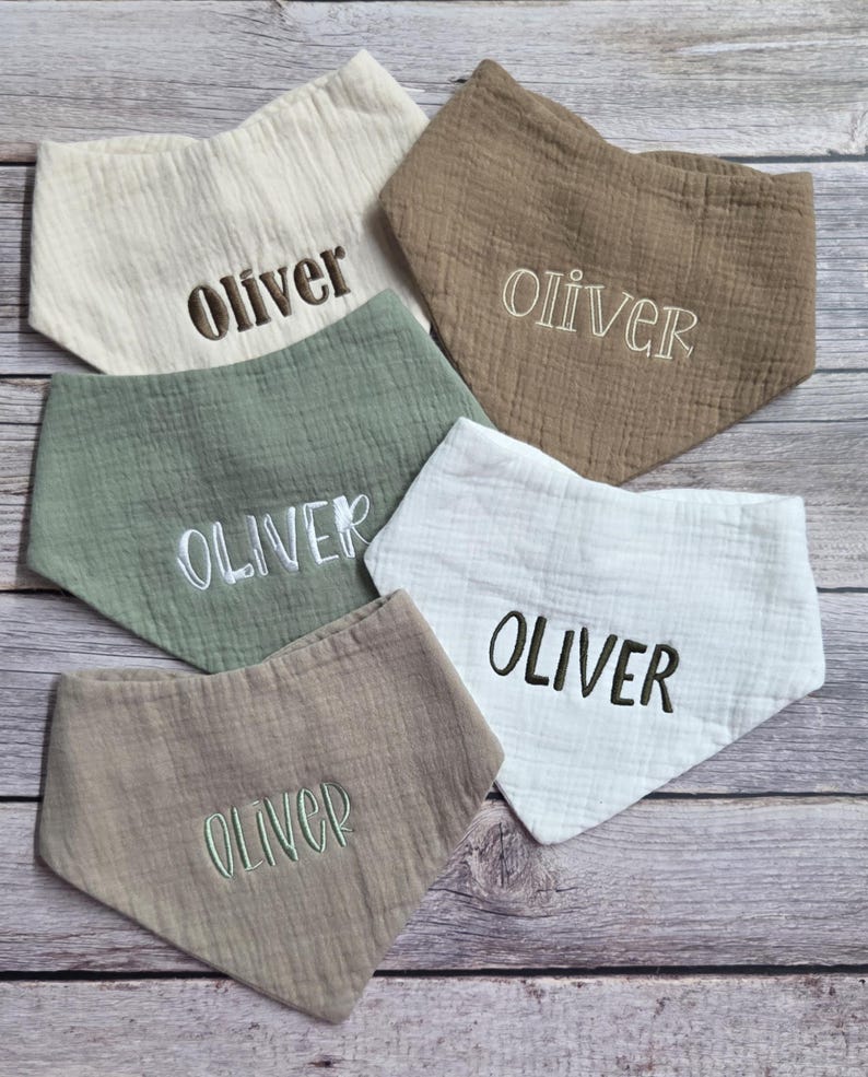May include: Five triangular baby bibs made of textured muslin fabric in cream, tan, sage green, and white. Each bib features the name Oliver embroidered in the center in various thread colors including dark brown, light tan, white, and green.
