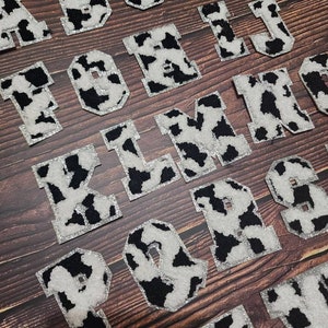 Peut inclure: Un ensemble de lettres en feutre &agrave; imprim&eacute; vache noir et blanc, de A &agrave; T, avec des accents de paillettes argent&eacute;es. Les lettres sont dispos&eacute;es en ordre alphab&eacute;tique sur une surface en bois.