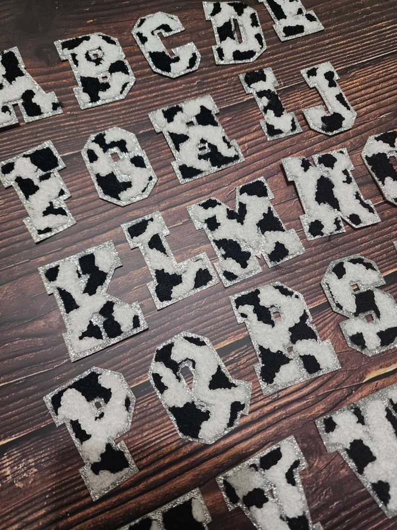 Peut inclure: Un ensemble de lettres en feutre &agrave; imprim&eacute; vache noir et blanc, de A &agrave; T, avec des accents de paillettes argent&eacute;es. Les lettres sont dispos&eacute;es en ordre alphab&eacute;tique sur une surface en bois.