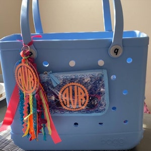 May include: A light blue tote bag with a clear zippered pouch. The pouch has a wave pattern and a monogram that says "AVR" in orange. The tote bag has a white number "9" on the side and a pink ribbon with a clear monogram that says "AVR" in orange.