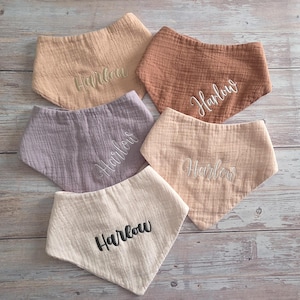 May include: Five triangular baby bibs made of textured muslin fabric in shades of beige, terracotta, mauve, and cream. Each bib features the embroidered name Harlow in either white or black script lettering, centered on the front of the fabric.