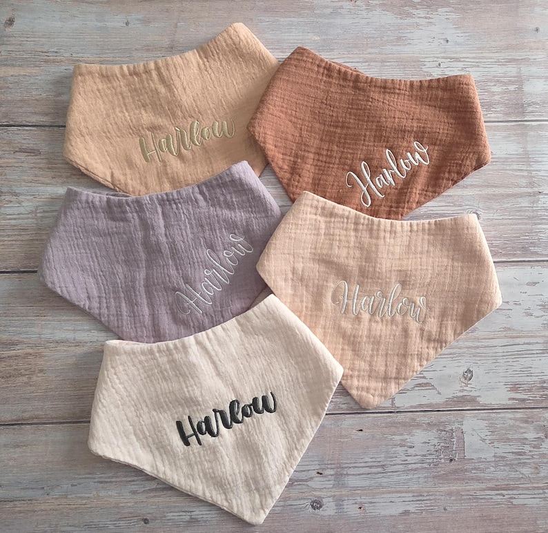May include: Five triangular baby bibs made of textured muslin fabric in shades of beige, terracotta, mauve, and cream. Each bib features the embroidered name Harlow in either white or black script lettering, centered on the front of the fabric.
