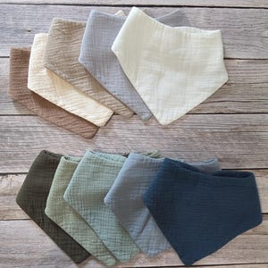 May include: A collection of triangular bibs made from textured muslin fabric in a variety of colors including cream, beige, light gray, dark gray, olive green, sage green, dusty blue, and navy blue, arranged in two overlapping rows.
