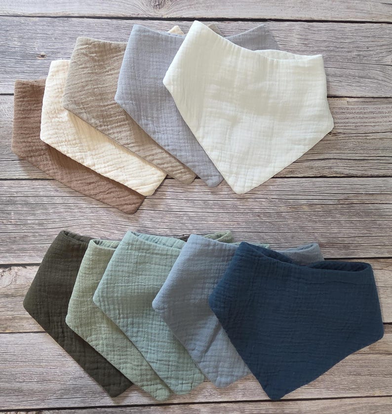 May include: A collection of triangular bibs made from textured muslin fabric in a variety of colors including cream, beige, light gray, dark gray, olive green, sage green, dusty blue, and navy blue, arranged in two overlapping rows.