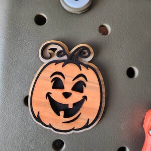 May include: A wooden pumpkin with a carved jack-o'-lantern face. The pumpkin is orange with black details and is attached to a green surface.