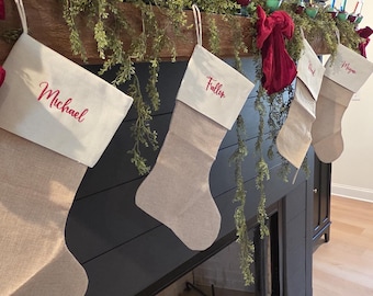 May include: A decorated fireplace mantel with Christmas stockings and vintage lights. The stockings are beige with red embroidered names. Greenery and red bows add festive touches. The mantel is dark, and the floor is light-colored wood.