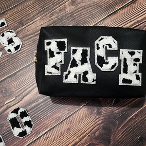 Peut inclure: Une trousse de maquillage noire &agrave; fermeture &eacute;clair avec le mot "FACE" &eacute;crit en lettres blanches et noires &agrave; imprim&eacute; vache.