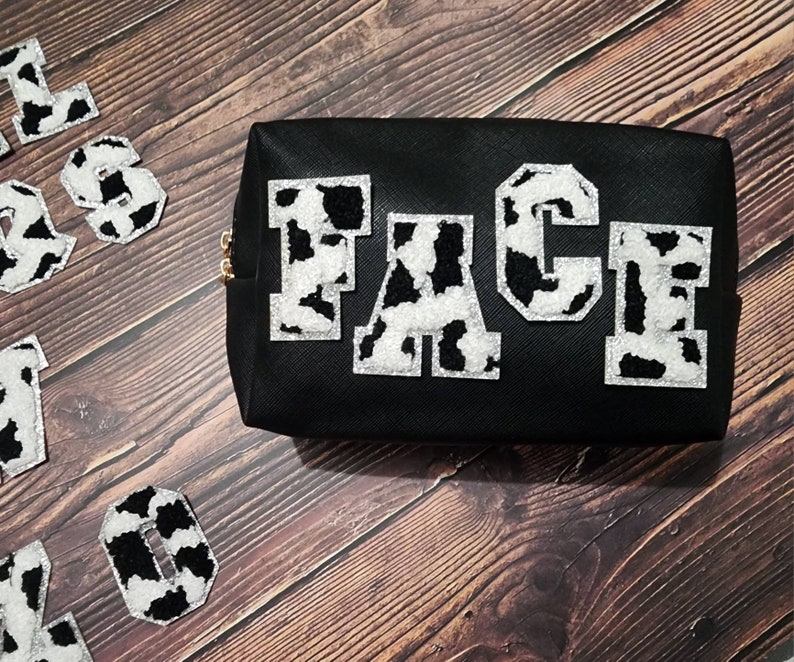 Peut inclure: Une trousse de maquillage noire &agrave; fermeture &eacute;clair avec le mot "FACE" &eacute;crit en lettres blanches et noires &agrave; imprim&eacute; vache.