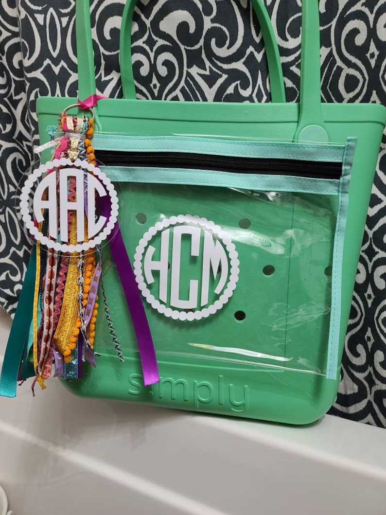 May include: A green clear tote bag with a zippered pocket and a white monogram that says "HCM". The bag has the word "simply" embossed on the bottom. A white monogram keychain with the letters "AAC" is attached to the bag.