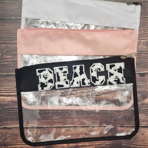 Peut inclure: Pochette zipp&eacute;e transparente en plastique avec un rabat en tissu noir qui dit "Beach" en lettres blanches avec un motif imprim&eacute; vache. La pochette est superpos&eacute;e &agrave; une pochette rose et une pochette blanche.