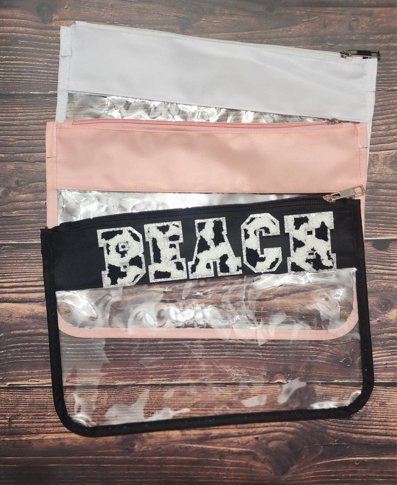 Peut inclure: Pochette zipp&eacute;e transparente en plastique avec un rabat en tissu noir qui dit "Beach" en lettres blanches avec un motif imprim&eacute; vache. La pochette est superpos&eacute;e &agrave; une pochette rose et une pochette blanche.