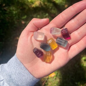 May include: A collection of small, colorful, rectangular crystals in various shades of purple, yellow, blue, and clear. The crystals are held in an open hand against a blurred green background.