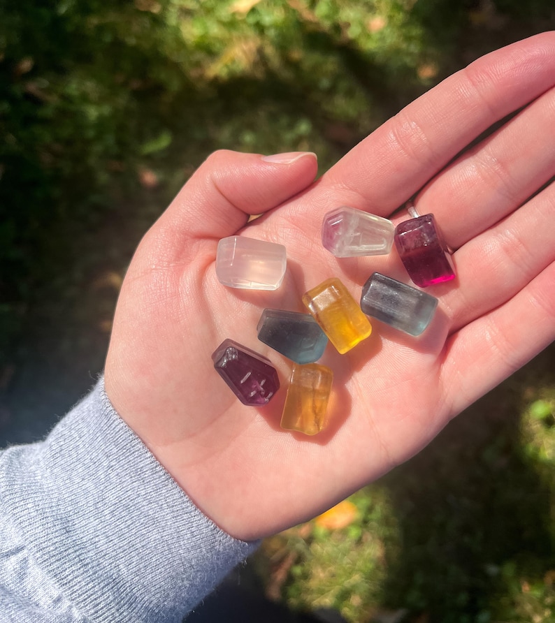 May include: A collection of small, colorful, rectangular crystals in various shades of purple, yellow, blue, and clear. The crystals are held in an open hand against a blurred green background.