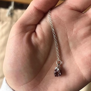 May include: A delicate silver chain necklace featuring a small, raw, deep red gemstone pendant. The pendant is secured with a silver-toned metal cap and a jump ring, suspended from a thin, polished silver-colored link chain that rests against the palm.