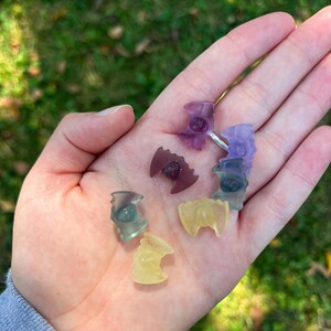 May include: A collection of small, colorful crystal bat figurines in shades of purple, green, and yellow. The figurines are held in a hand against a blurred green background. The crystals have a translucent quality.