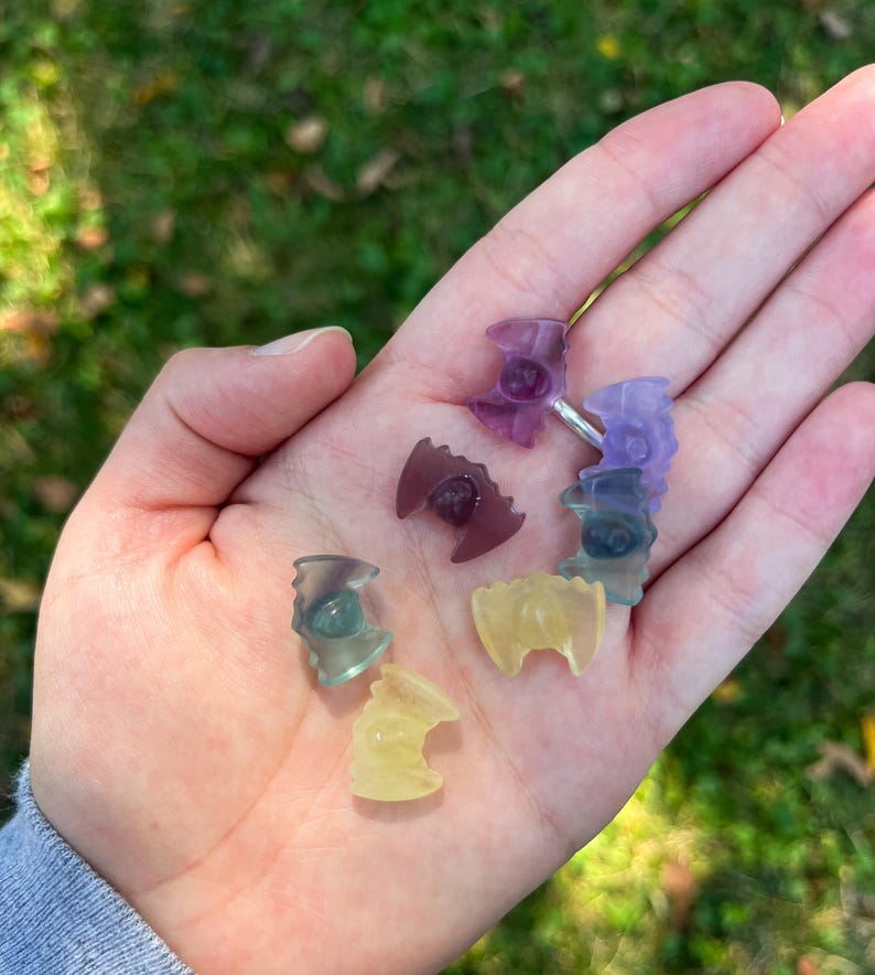 May include: A collection of small, colorful crystal bat figurines in shades of purple, green, and yellow. The figurines are held in a hand against a blurred green background. The crystals have a translucent quality.