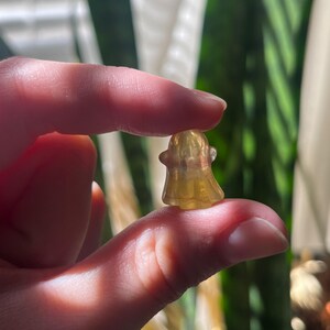 May include: A small, translucent yellow ghost figurine. The ghost has a simple, rounded shape with a small head and a flowing body. The figurine is held between two fingers, with a blurred green background.